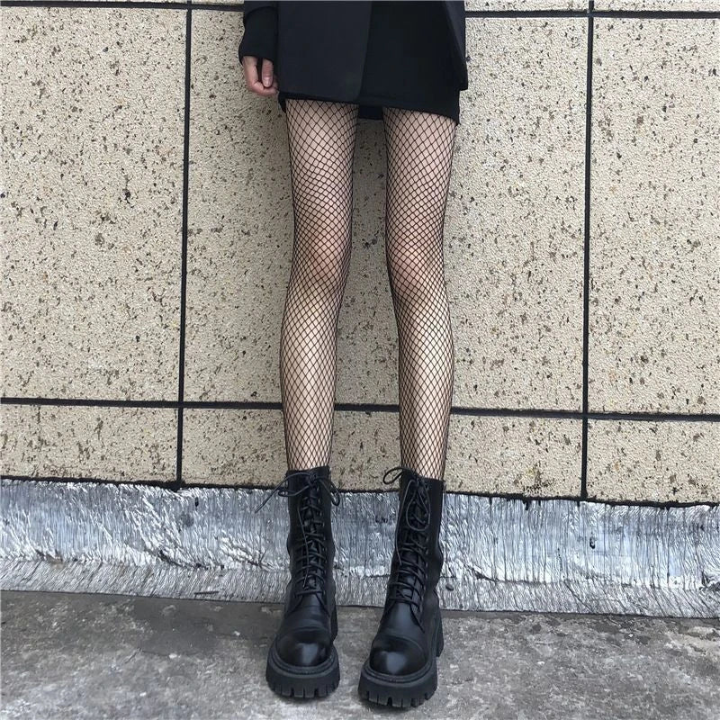 Person wearing black fishnet stockings and black combat boots against a tiled wall.